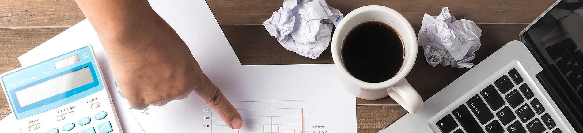 A person pointing to a bar graph with a calculator to the left, and a laptop computer and cup of coffee to the right.