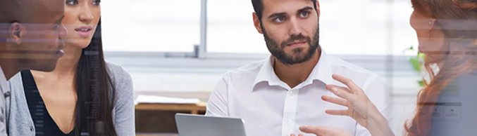 A small group of young colleagues sits around a laptop in an office setting, actively engaged in discussion.