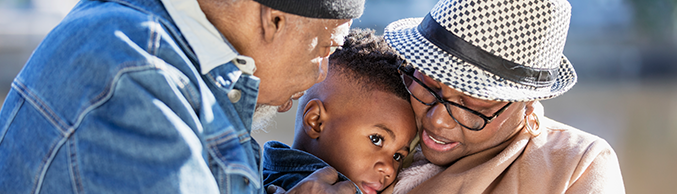 A 3 year old boy in the park with his grandparents on a sunny autumn day. His grandmother is holding him in her arms. His grandfather is looking at him, with his hand on the boy's shoulder.