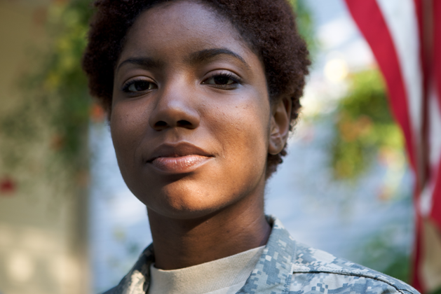 A young veteran dressed in camouflage focuses on the camera while an American flag hangs in the background